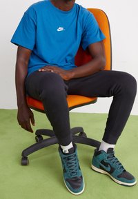 Modré bavlněné tričko s bílým logem Nike, černé tepláky a tyrkysové vysoké tenisky na židli s oranžovým opěradlem na zelené podlaze.