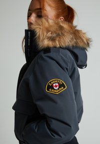 Tumši zili izolēta jaka ar mākslīgā pūka kapuci. Iezīmēta ar logo uz piedurknes un izgatavota no plānām, izturīgām materiālām.