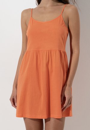 Vestito estivo arancione senza maniche con spalline sottili e vita arricciata indossato da una persona in piedi su uno sfondo semplice.