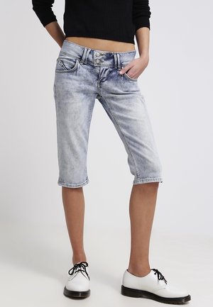 Personne portant un short en denim bleu clair longueur genou, un pull court noir et des chaussures blanches à lacets. Mains dans les poches.