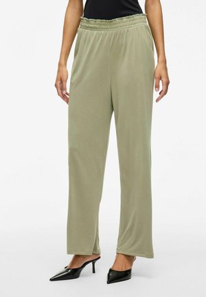 Pantalones de pierna ancha en verde oliva claro con cintura elástica fruncida, hechos de una tela suave, combinados con tacones negros de punta. 