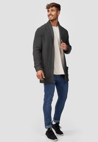 Cardigan in maglia di carbone con texture a coste, apertura frontale e tasche, abbinato a una t-shirt chiara e jeans blu, e sneaker nere.