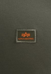 Mörk olivfärgad tyg med en texturerad yta har en rektangulär svart lapp sydd med "ALPHA INDUSTRIES" i orange, vilket framhäver varumärkeslogotypen.
