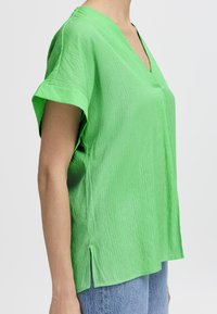 Top verde a maniche corte con scollatura a V, realizzato in un tessuto testurizzato, con spacchi laterali e una vestibilità rilassata. Indossato sopra jeans in denim blu.