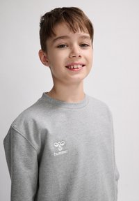 Felpa grigia con collo a giro. Presenta un piccolo logo bianco "hummel" sul lato sinistro del petto. Il materiale sembra morbido e liscio.