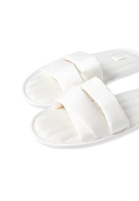 Pantofole bianche in tessuto, con una texture trapuntata, dotate di due larghe cinghie imbottite sulla parte superiore e una suola morbida e imbottita.