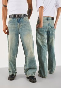 Vaqueros de color azul claro, de corte holgado, con un lavado desgastado, que presentan bolsillos profundos, costuras visibles y un parche de cuero en la cintura. Incluye cinturón negro.