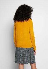 Gul stickad tröja med långa ärmar och rundad nederkant, i par med en mönstrad kjol som visar cirkulära motiv i blått och vitt.