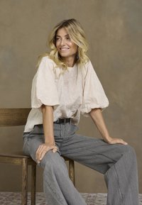 Vaaleanpunainen paita, jossa on puffatut hihat ja kukkakirjonta, yhdistettynä korkean vyötärön raidallisiin kangashousuihin. Puiset tuolit neutraalilla taustalla.