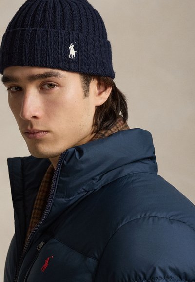 Mand iført en navy strikket hue og navy dunjakke med et rødt logo, der kigger mod kameraet mod en neutral baggrund.