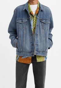 Blå denimjacka med två bröstfickor, knappstängning och en avslappnad passform, lager på en flerfärgad mönstrad skjorta.