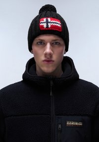 Berretto di lana nero con design della bandiera norvegese rosso, bianco e blu e un pompon sopra. Indossato con una giacca in pile nera con logo.