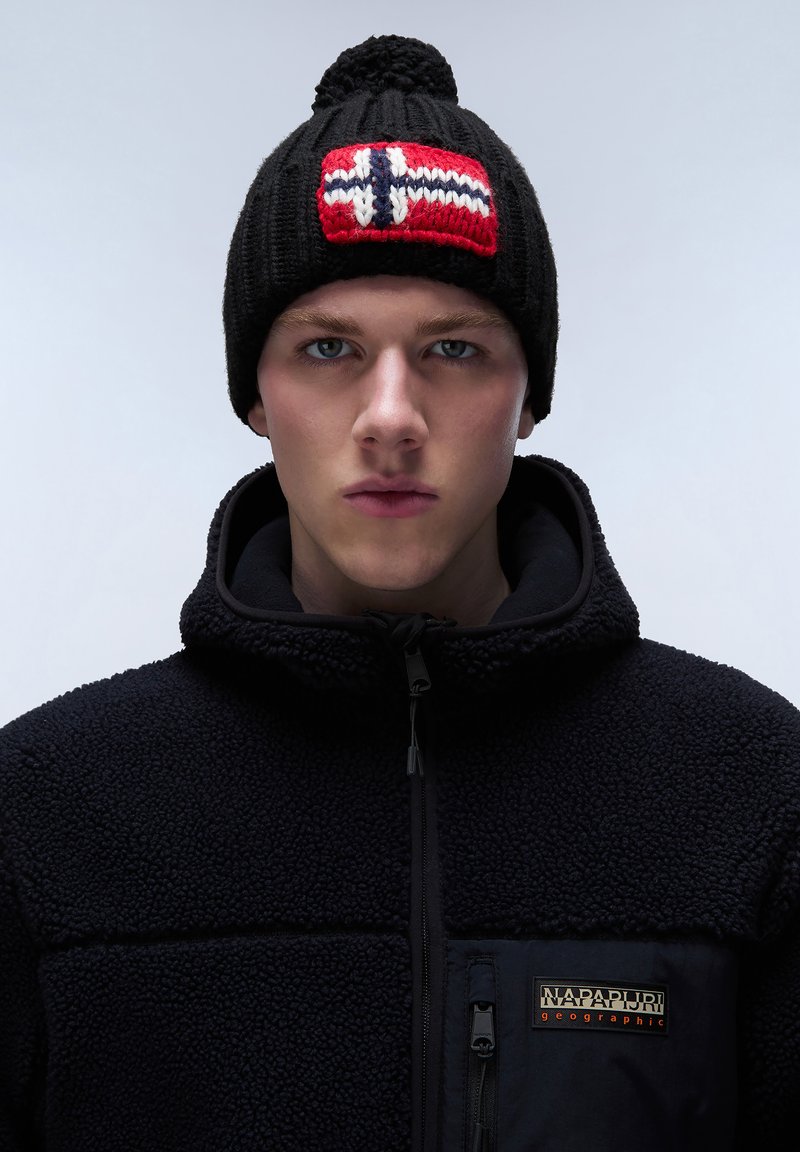 Berretto di lana nero con design della bandiera norvegese rosso, bianco e blu e un pompon sopra. Indossato con una giacca in pile nera con logo.