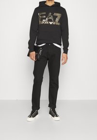 Svart hoodie med stort guld "EA7 Emporio Armani"-logotyp, matchad med svarta byxor och svarta högskurna sneakers med vit sula.
