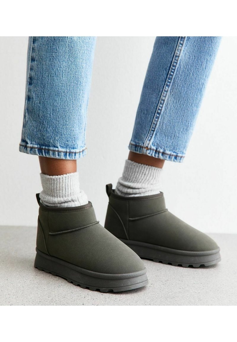Personne portant des bottines grises avec des chaussettes blanches épaisses et un jean bleu clair, debout sur un sol clair sur un fond blanc.
