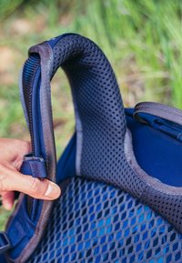 Bretelle de sac à dos en maille bleue avec un bord rembourré. Présente des trous de ventilation pour la respirabilité. Main tenant la sangle avec une boucle réglable visible.
