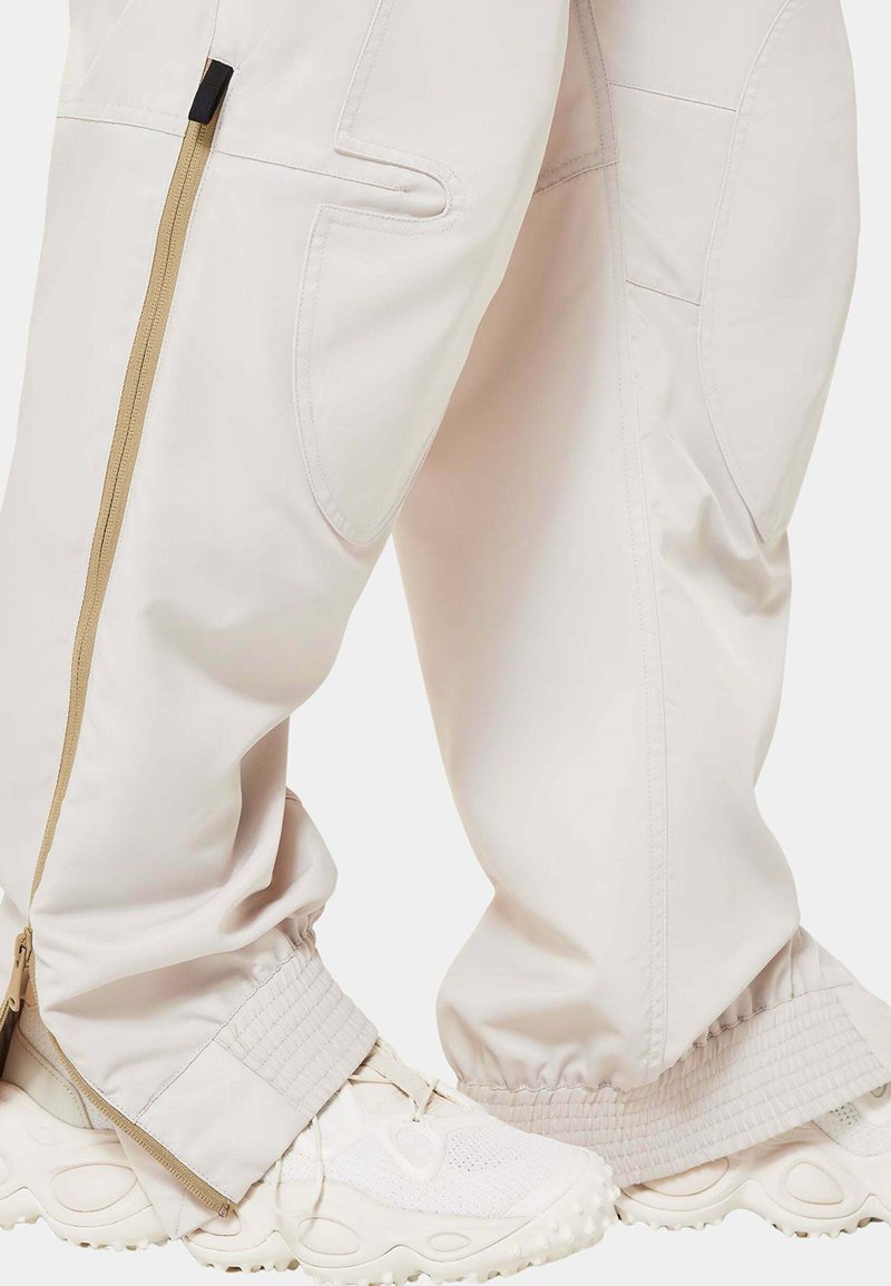 Helle beigefarbene Hosen mit Reißverschlusstaschen und elastischen Bündchen, kombiniert mit weißen strukturierten Turnschuhen mit einer klobigen Sohle.