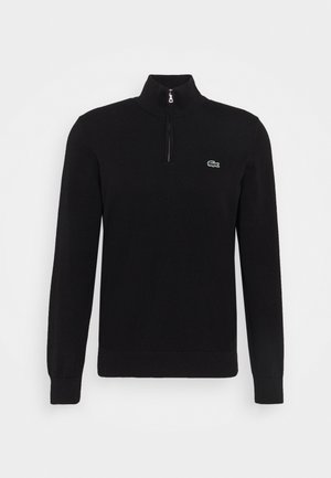 Pull en tricot noir avec un col haut et une fermeture éclair mi-hauteur. Présente un logo Lacoste vert sur la poitrine. Manches longues et poignets côtelés.