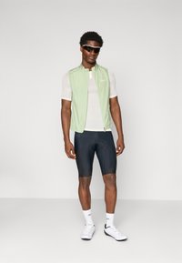 Hellgrüne ärmellose Radweste über einem weißen Kurzarmhemd, kombiniert mit schwarzen Radshorts. Das Model trägt eine Sonnenbrille und weiße Schuhe.