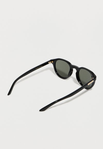 Μαύρα στρογγυλά γυαλιά ηλίου με σκούρους φακούς και χρυσές λεπτομέρειες στις μεντεσέδες, τοποθετημένα σε λευκή επιφάνεια.