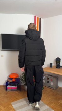 Black puffer jacket with a detachable hood, featuring a quilted design and elastic cuffs, paired with loose black pants and grey sneakers.