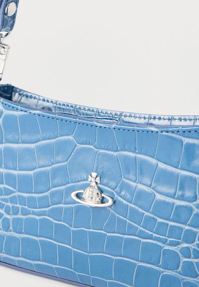 Μπλε τσάντα χειρός με υφή κροκοδείλου, λογότυπο ασημένιας σφαίρας και μερικώς ορατό λουράκι σε απλό φωτεινό φόντο.
