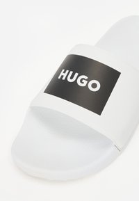 Bijela sandala za klizanje s teksturiranom potplatom i širokom trakom koja ima crni pravokutni panel s natpisom "HUGO" u bijeloj boji.