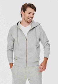 Szara bluza z kapturem na zamek błyskawiczny, z przodu, z ściągaczami na rękawach i luźnym krojem. Noszona na białej koszulce, posiada kaptur i gładką fakturę.