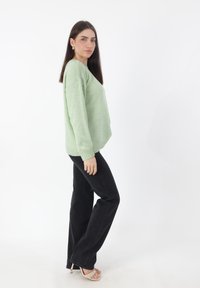 Hellgrüner Strickpullover mit lässiger Passform und gerippten Bündchen, kombiniert mit schwarzen Schlagjeans und beigen, hochhackigen Sandalen, vor einem einfarbigen Hintergrund.