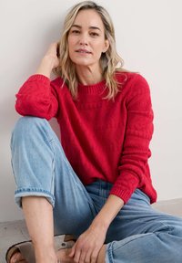Maglione rosso lavorato a maglia con motivi testurizzati, scollatura rotonda e polsini a costine, abbinato a jeans azzurri arrotolati e sandali beige.