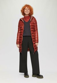 Rode pufferjas met een gequilte textuur, ritssluiting en capuchon. Gepaard met een donkere trui en wijde zwarte broek. Chunky zwarte laarzen.
