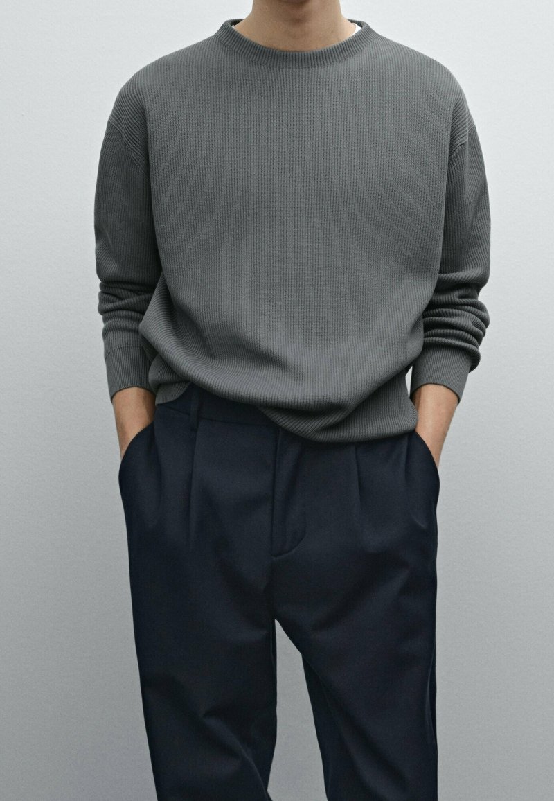 Grauer gerippter Pullover mit Rundhalsausschnitt, kombiniert mit dunkelblauen Hosen mit Seitentaschen und lockerer Passform.