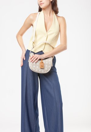 Beige geprinte schoudertas met een tan leren label, met een ronde vorm en een witte schouderband, gedragen met een gele top en blauwe broek.