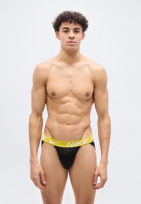 Νέος μυώδης άνδρας με σγουρά μαλλιά, φοράει μαύρα εσώρουχα με κίτρινη μέση Nike, στέκεται μπροστά σε απλό φωτεινό φόντο.