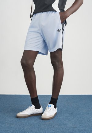 Pantalones cortos deportivos azul claro con rayas laterales negras y un logo de adidas, combinados con zapatillas blancas con detalles azules y suelas marrones.