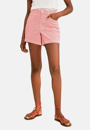 Person iført røde og hvide gingham-shorts, hvid top og røde sandaler med remme og flade såler, stående med den ene hånd i lommen.