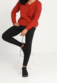 Pull rouge en maille torsadée avec col en V ; associé à des leggings noirs rayés et des baskets noires. La tenue présente une coupe décontractée et des motifs texturés.
