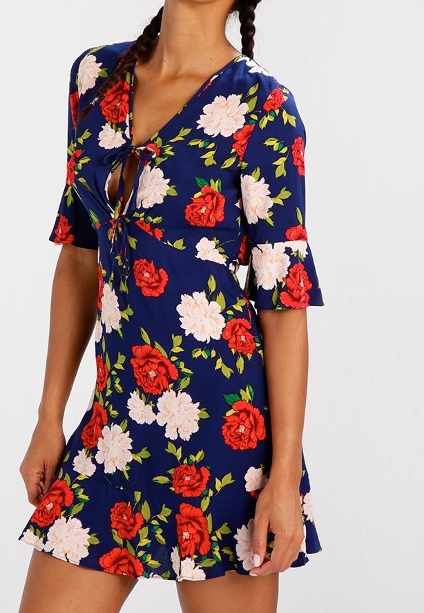 Femme portant une robe courte bleue à fleurs rouges et blanches avec un col en trou de serrure noué à l'avant, debout devant un fond uni.