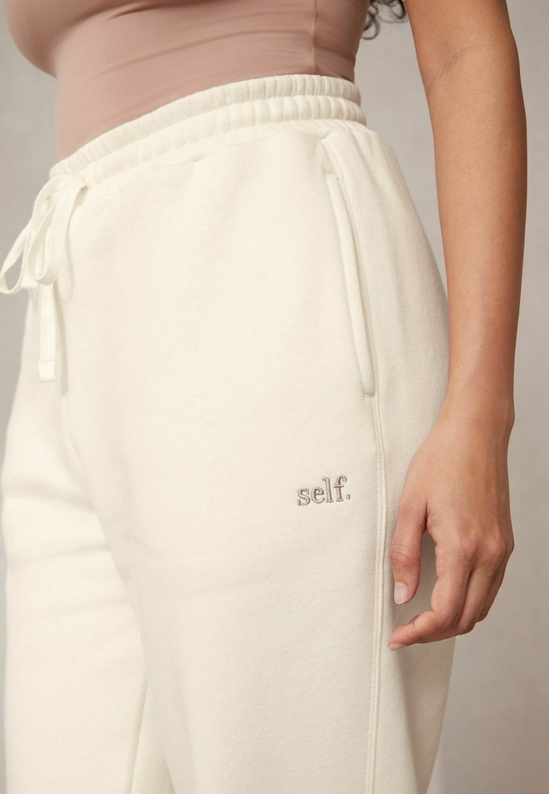 Cremefarbene Sweatpants aus weichem Stoff mit einem Kordelzugbund, versehen mit einem gestickten Logo "self." an der Seite.