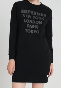 Černé šaty s dlouhým rukávem, na kterých je stříbrný nápis: "SUPERDRY," "NEW YORK," "LONDON," "PARIS," "TOKYO." Měkká tkanina, volný střih.
