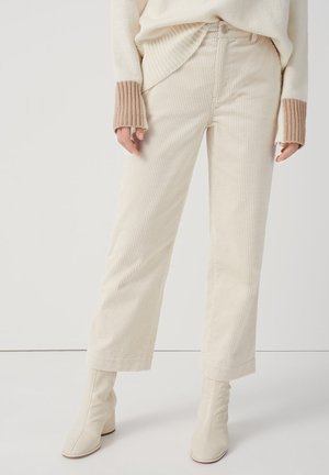 Frau trägt breite Cordhosen in Creme, cremefarbenen Pullover mit braunen, gerippten Bündchen und cremefarbene Stiefeletten, steht auf weißem Boden.