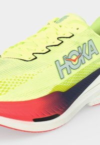 Světle žlutá a červená běžecká bota Hoka s černými akcenty na tkaničkách a bílou podrážkou, zobrazena zblízka na jednoduchém pozadí.