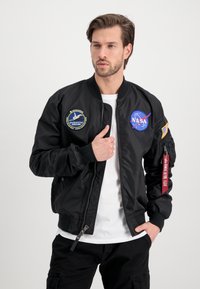 Black bomber jacket with NASA patches on chest and sleeve, ribbed cuffs, and zipper detail. Smooth fabric, slightly fitted design.