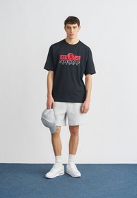 Μαύρο γραφικό T-shirt με κόκκινο και λευκό λογότυπο "Jordan", γκρι σορτς, λευκά αθλητικά παπούτσια, γκρι καπέλο κρατημένο στο χέρι, στέκεται σε μπλε χαλί.