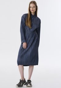 Vestido camisero de mezclilla oscura con rayas verticales, mangas largas, cierre frontal de botones, cintura ajustada y largo hasta la rodilla. Acentuado con detalles cosidos.