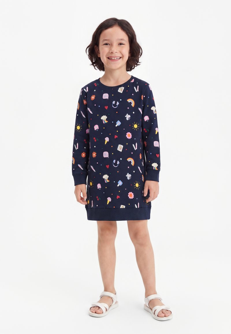 Enfant souriant aux cheveux courts et foncés, portant une robe bleu marine décorée de soleils colorés, d'arcs-en-ciel, de fleurs et de cœurs, ainsi que des sandales blanches.