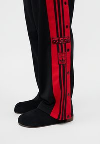Schwarze Hose mit roten Seitenstreifen und schwarzem Adidas-Logo, Druckknöpfe entlang des roten Streifens, getragen mit schwarzen Slippern.