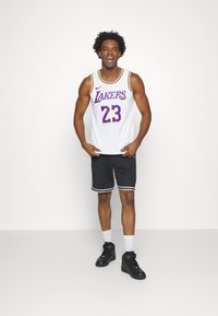 Bijela košarkaška majica s ljubičastim detaljima, natpis "Lakers" i broj 23. Uparen s crnim kratkim hlačama s bijelim obrubom i crnim tenisicama.