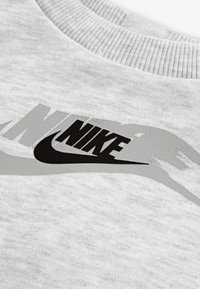 Hellgrauer Sweatshirt mit geripptem Halsausschnitt, das ein auffälliges schwarzes Nike-Logo und ein dezentes graues Grafikdesign aufweist. Weicher, strukturierter Stoff.