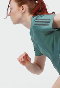 Red-haired woman in teal athletic shirt bends forward, appearing focused and poised to run or engage in physical activity.